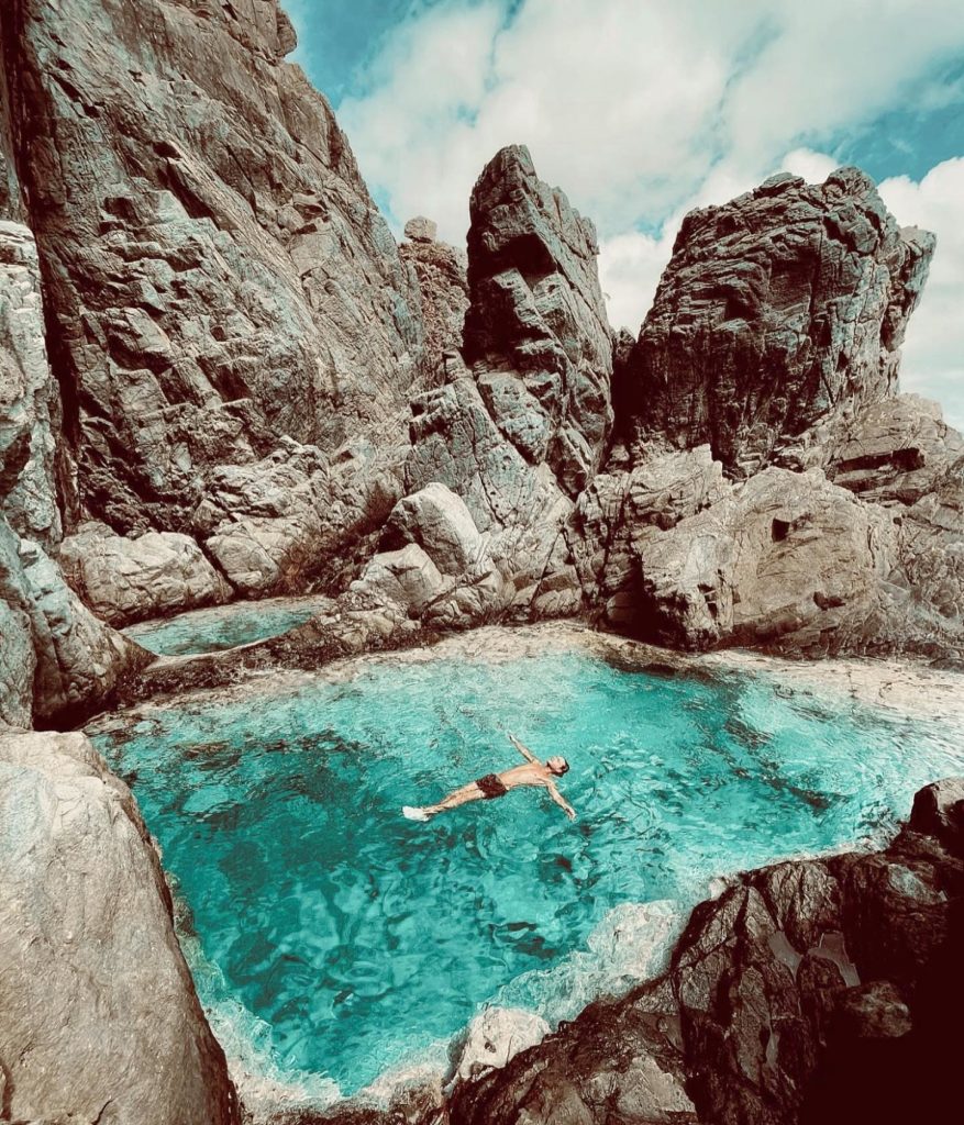 Man floating on his back in bright blue water surrounded by rocks.