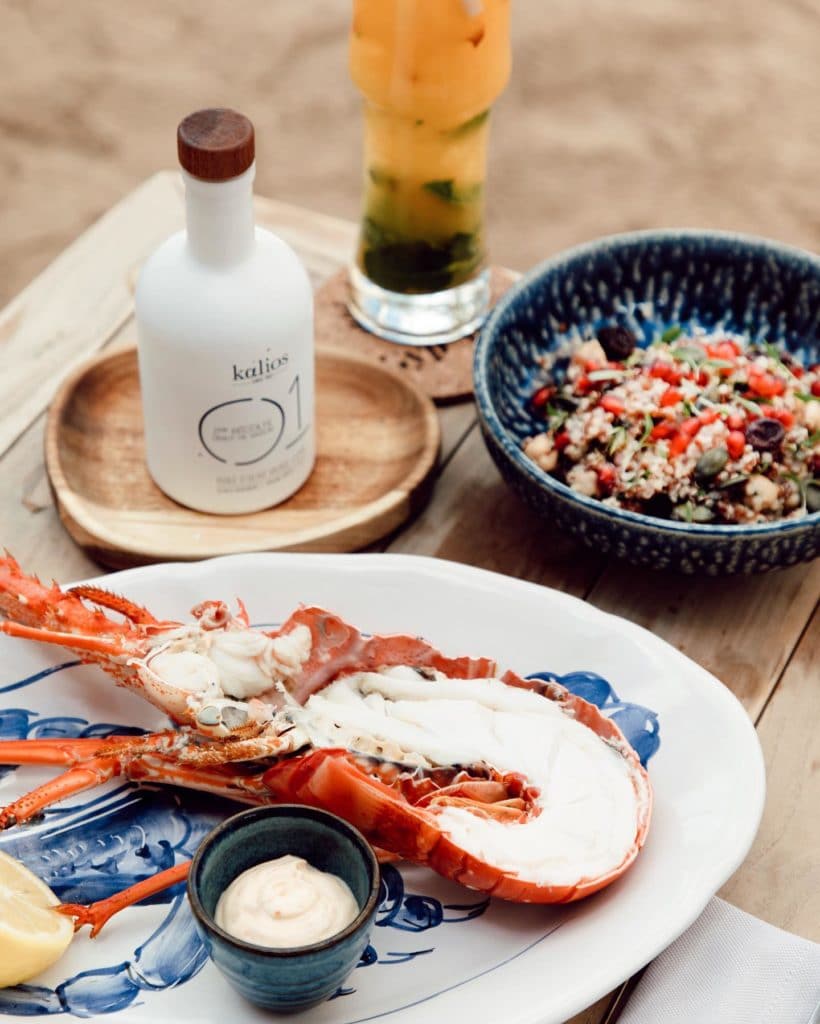Halved lobster on ceramic blue and white plate on top of a wooden table on a beach surrounded by Mediterranean quinoa salad, olive oil, and a tropical cocktail in a glass.