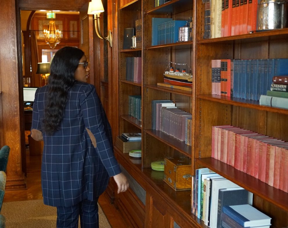 Woman in a navy, plaid suit walking in a library in a hotel. The library has many books in the wooden bookcase.