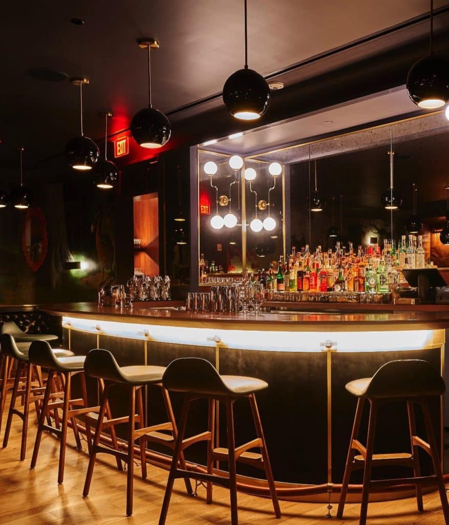 Speakeasy-style bar with various bottles on the bar and bar stools surrounding the curved bar area.