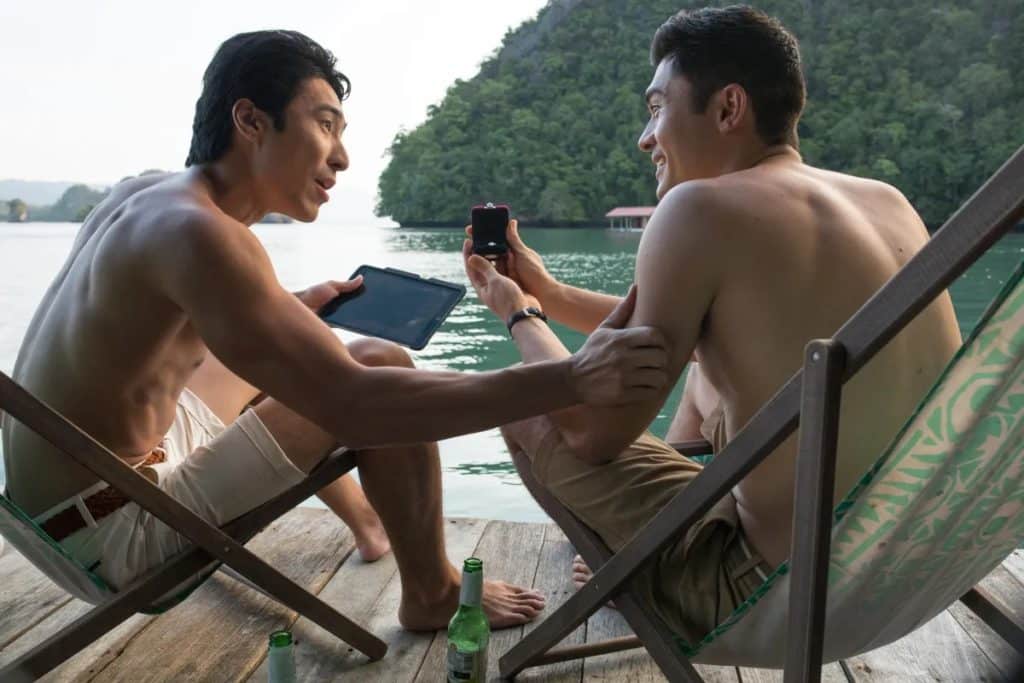 Scene from the movie Crazy Rich Asians with Henry Golding sitting by the water and showing his friend a ring.