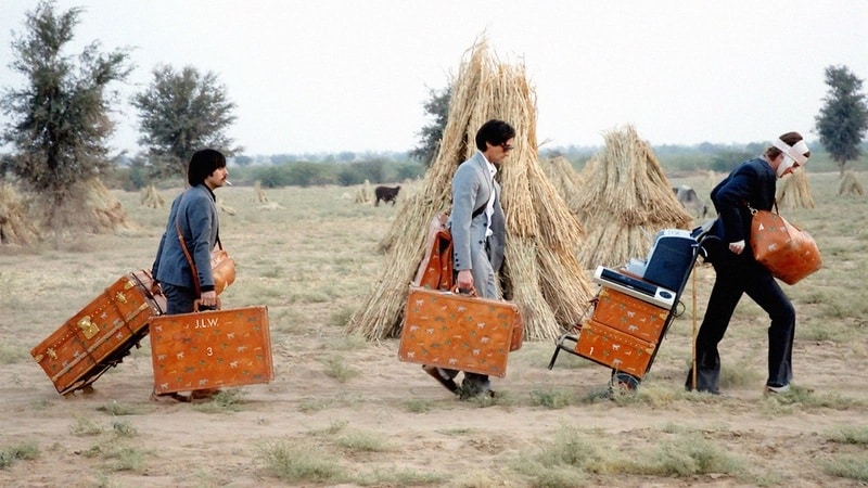 Scene from The Darjeeling Limited movie where three men are carrying their luggage across a grassy field.