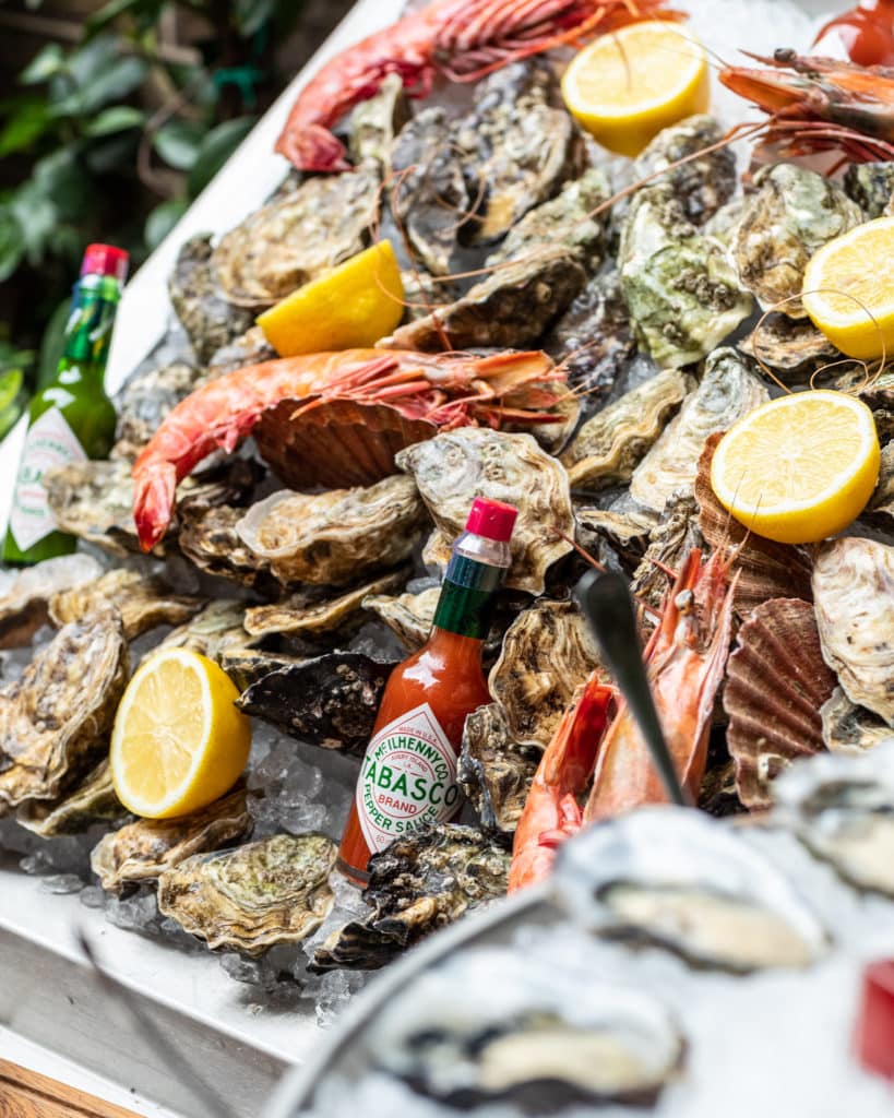 Various types of shellfish, including oysters, on a white platter with ice. The seafood is served with lemon and Tabasco sauce.