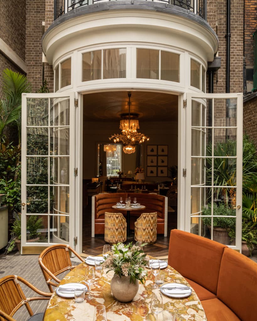 Elegant outdoor dining setting. There is a table at the center of the area overlooking two large doors and the indoor dining setting. The table and chairs are an orange color and are near a brick building.