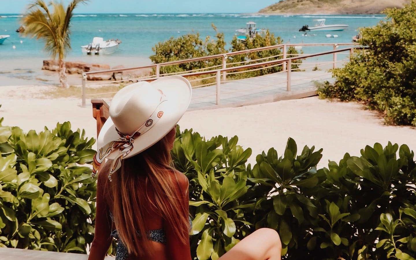 Woman in bikini and white straw wide-brimmed hat sitting on edge of lounge chair with back to camera looking out at white sandy beach, palm trees, and turquoise ocean.